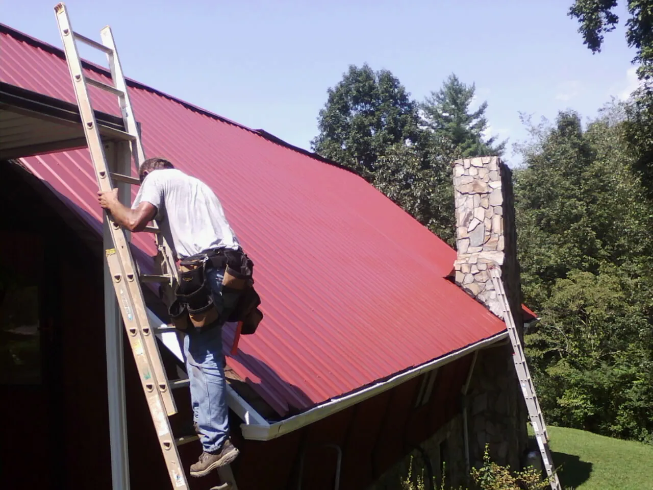 Roofer on a ladder beside a red metal roof and stone chimney — Soffit & Fascia Repair by Roofing Brothers in Luthersville